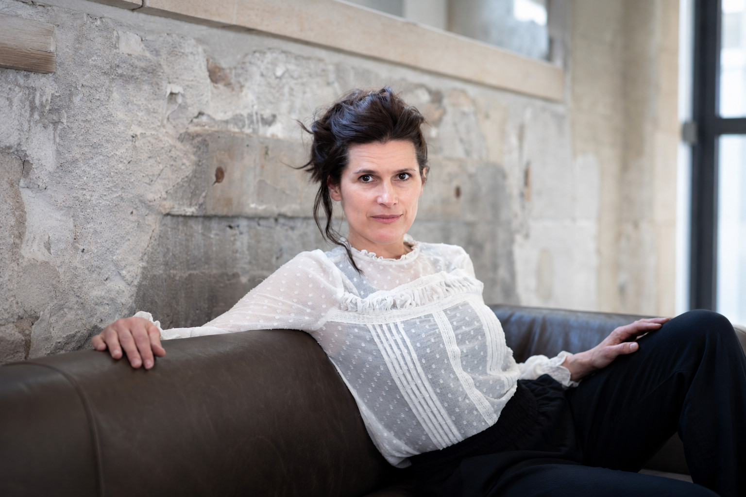 Margarete Lamprecht in weißer Spitzenbluse auf einem Sofa vor einer Betonwand